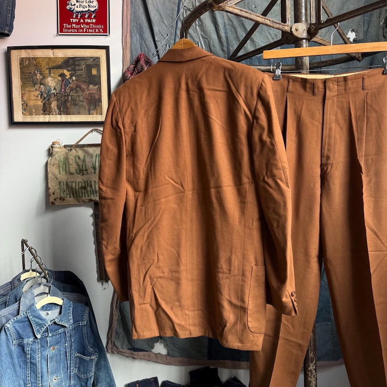 May include: A solid brown suit consisting of a single-breasted blazer with a notched lapel and matching trousers with a pleated front. The blazer features long sleeves with buttoned cuffs and a straight hem, while the trousers have a high waist.