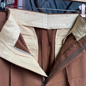 May include: A pair of brown trousers featuring a high-waisted design with a cream-colored interior lining. The garment includes a metal zipper fly closure and a structured waistband with visible stitching, suspended from a metal clothing hanger.