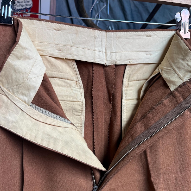 May include: A pair of brown trousers featuring a high-waisted design with a cream-colored interior lining. The garment includes a metal zipper fly closure and a structured waistband with visible stitching, suspended from a metal clothing hanger.