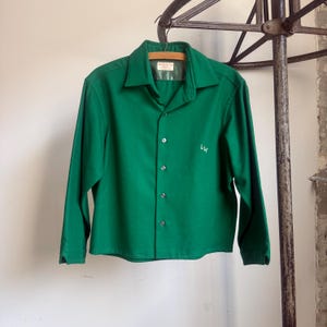 May include: A vintage emerald green button-down shirt with long sleeves, a collar, and a small embroidered design on the front. The shirt is hanging on a wooden hanger. The shirt is made of a soft fabric.