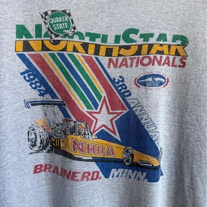 May include: A heather gray t-shirt featuring a vintage 1984 North Star Nationals graphic. The design includes a yellow NHRA dragster, a Quaker State flag, and text reading 3rd Annual Brainerd, Minn. The graphic uses green, yellow, red, and blue.