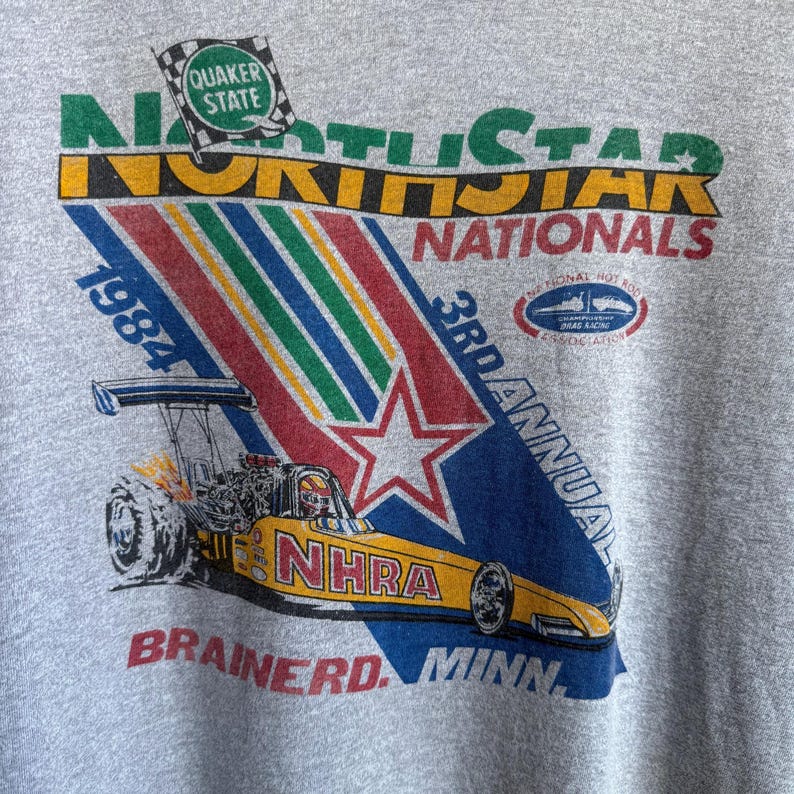 May include: A heather gray t-shirt featuring a vintage 1984 North Star Nationals graphic. The design includes a yellow NHRA dragster, a Quaker State flag, and text reading 3rd Annual Brainerd, Minn. The graphic uses green, yellow, red, and blue.