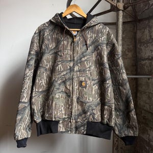 Puede incluir: Chaqueta con capucha Carhartt con un patr&oacute;n de camuflaje marr&oacute;n, verde y beige. La chaqueta tiene una cremallera completa, pu&ntilde;os y dobladillo de canal&eacute; negros y un forro de capucha negro. Un peque&ntilde;o parche con el logotipo de Carhartt est&aacute; presente.