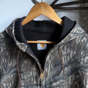 Puede incluir: Chaqueta con capucha Carhartt con estampado de camuflaje, colgada de una percha de madera. La chaqueta tiene un forro interior negro, una cremallera completa y cordones marrones. El logotipo de Carhartt es visible en el pecho.