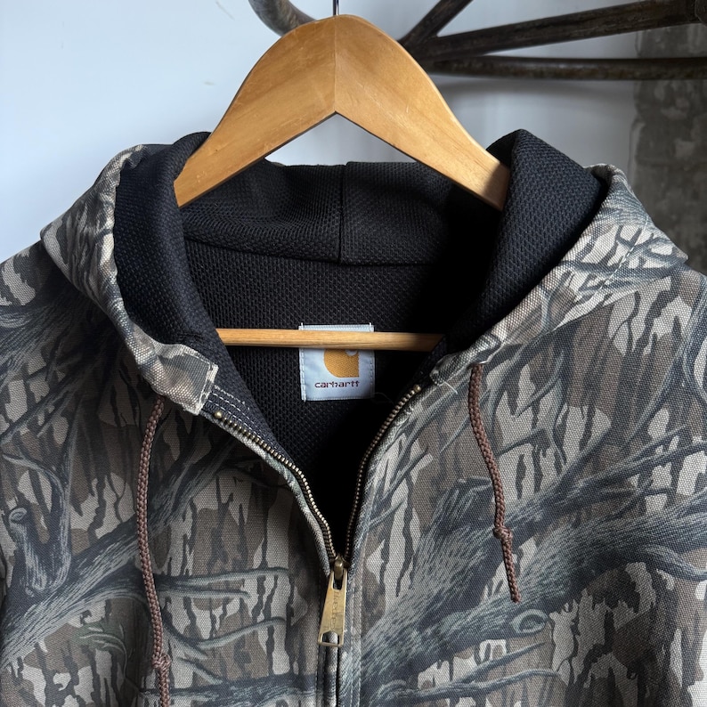 Puede incluir: Chaqueta con capucha Carhartt con estampado de camuflaje, colgada de una percha de madera. La chaqueta tiene un forro interior negro, una cremallera completa y cordones marrones. El logotipo de Carhartt es visible en el pecho.