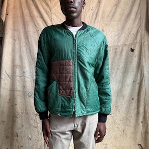 May include: A quilted green bomber jacket featuring a central front zipper, a black ribbed collar, and black ribbed cuffs. The jacket includes a contrasting brown quilted patch on the right chest area and a white shirt visible underneath.