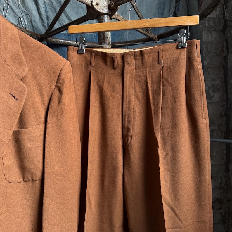May include: A pair of brown trousers with a pleated front, belt loops, and a side pocket, hanging on a wooden hanger next to a matching brown blazer. The trousers feature a straight-leg cut and a smooth, solid-colored fabric construction.