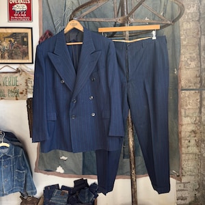 May include: A dark blue double-breasted suit featuring a subtle vertical pinstripe pattern. The set includes a blazer with wide peak lapels and matching trousers with cuffed hems, both displayed on wooden hangers against a fabric backdrop.