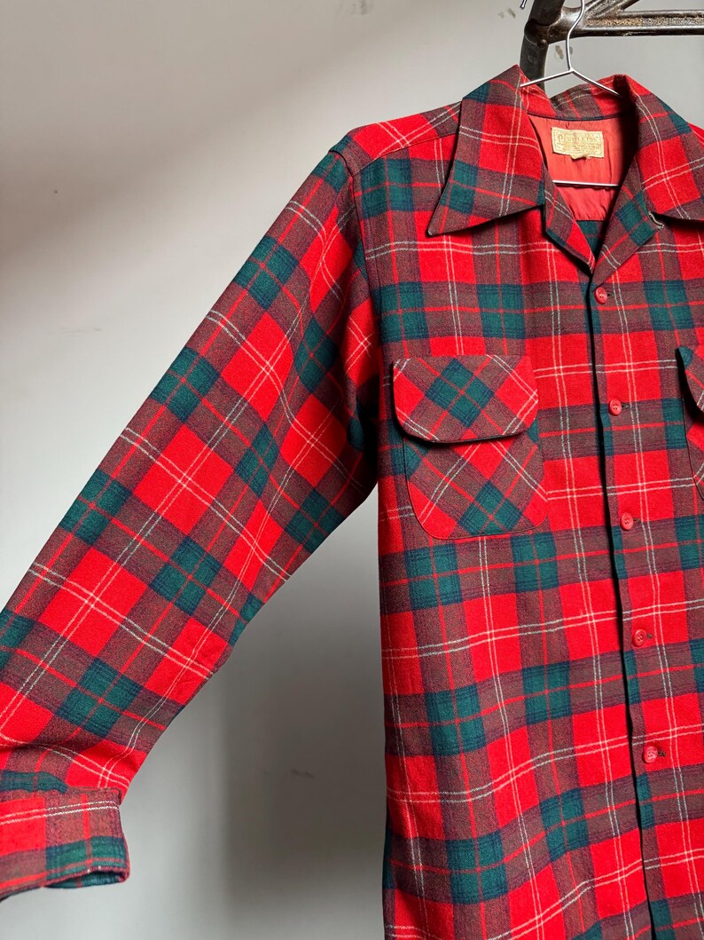 K&ouml;nnte beinhalten: Ein rot, gr&uuml;n und braun kariertes Button-Down-Hemd mit langen &Auml;rmeln. Das Hemd hat zwei Brusttaschen und einen Kragen. Der Stoff scheint ein mittelschweres Material zu sein, das f&uuml;r k&uuml;hleres Wetter geeignet ist.