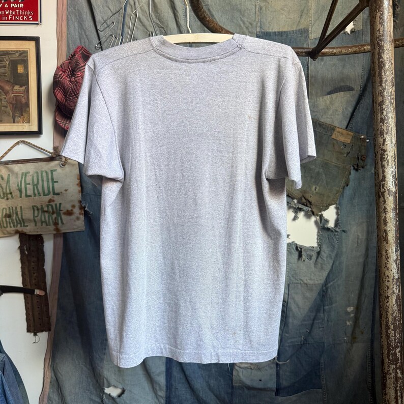 May include: A plain light gray short-sleeve t-shirt with a crew neckline. The garment is displayed hanging on a white hanger, showing a simple, solid-colored design with a standard fit, short sleeves, and a straight hemline at the bottom edge.