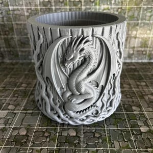 May include: A gray cylindrical container with a detailed relief of a dragon with outstretched wings. The container has a textured, tree-like appearance with intricate carvings. The dragon is centered on the container.