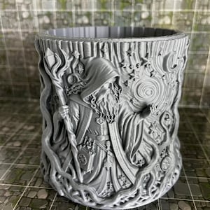 May include: A gray, cylindrical container with a detailed relief of a wizard holding a staff. The wizard is surrounded by stars, vines, and a crescent moon. The container is likely 3D-printed and suitable for holding pens or small items.