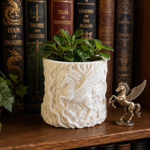 May include: A white cylindrical planter featuring a relief sculpture of a winged horse, stars, and a crescent moon, containing a small green plant. Beside the planter stands a small, metallic bronze-colored figurine of a winged horse.
