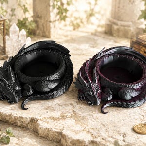 May include: Two dragon-shaped candle holders. One is black, the other is black with burgundy accents. The dragons are curled around a circular bowl, with detailed scales and horns. The candle holders are on a stone surface.