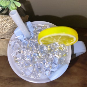 Pu&ograve; includere: Una tazza blu chiaro con manico, piena di ghiaccio e una fetta di limone. Una cannuccia bianca sporge dalla tazza.