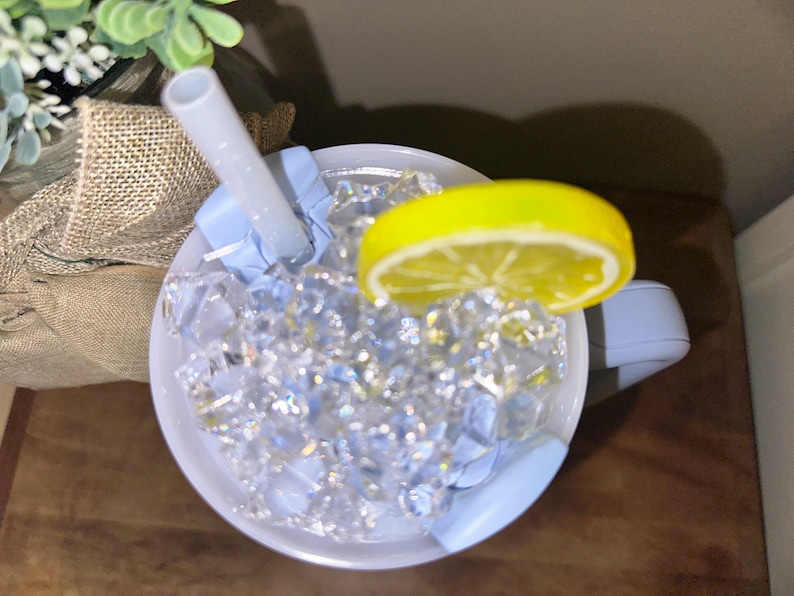 Pu&ograve; includere: Una tazza blu chiaro con manico, piena di ghiaccio e una fetta di limone. Una cannuccia bianca sporge dalla tazza.