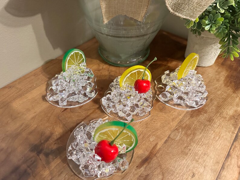 Pu&ograve; includere: Quattro sottobicchieri in acrilico trasparente con un design decorativo di cubetti di ghiaccio, una ciliegia e una fetta di limone o lime.