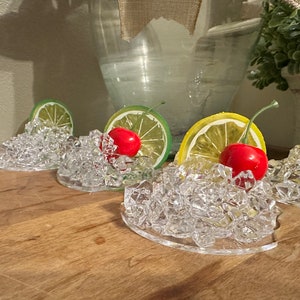 Pu&ograve; includere: Quattro porta cubetti di ghiaccio in plastica trasparente con una fetta di lime o limone e una ciliegia rossa in cima. I porta cubetti sono su una superficie di legno.