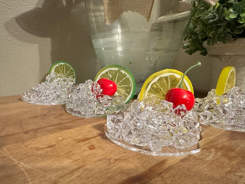 Pu&ograve; includere: Quattro porta cubetti di ghiaccio in plastica trasparente con una fetta di lime o limone e una ciliegia rossa in cima. I porta cubetti sono su una superficie di legno.