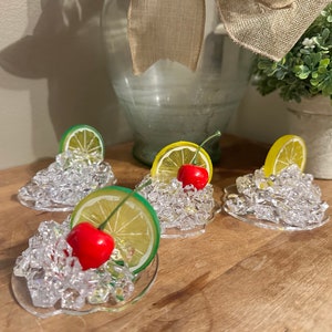 Pu&ograve; includere: Quattro sottobicchieri in vetro trasparente con una base di ghiaccio simulato, ognuno sormontato da una fetta di limone o lime e una ciliegia rossa.