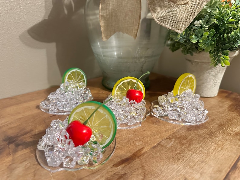 Pu&ograve; includere: Quattro sottobicchieri in vetro trasparente con una base di ghiaccio simulato, ognuno sormontato da una fetta di limone o lime e una ciliegia rossa.