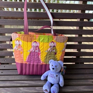 May include: A quilted tote bag with a purple and yellow floral pattern featuring women in dresses. The bag has a purple leather bottom and two straps. A purple teddy bear with the text "Got Wine?" is sitting in front of the bag.