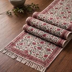 May include: A rectangular table runner featuring a cream-coloured base with a repeating floral pattern in deep red and dark green. The fabric has a decorative border and is finished with off-white fringe tassels along the short ends of the runner.