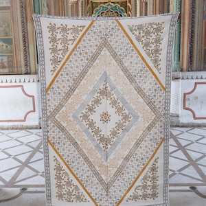 Può includere: Tessuto color crema con motivo a rombi. Il design presenta motivi floreali e geometrici nei toni dell'oro, del grigio e del marrone. Il bordo è grigio scuro con un motivo ripetuto. Il tessuto è esposto in una stanza con dettagli architettonici ornati.