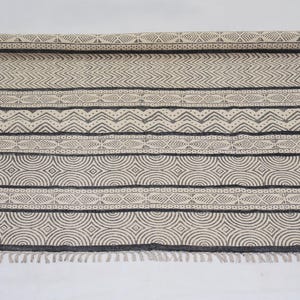 May include: Beige and gray patterned rug with a rolled-up section. The rug features geometric designs, including stripes, chevrons, and circles. The edges have a fringe detail. A decorative floor covering.