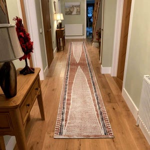 May include: A long, narrow runner rug featuring a geometric pattern in shades of terracotta, cream, and dark charcoal. The design includes elongated triangular shapes and intricate border details, laid out on a light-coloured wooden floor in a hallway.