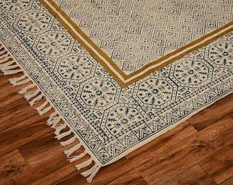 May include: A white cotton rug with a blue and gold geometric pattern. The rug has a fringe border and is made with a hand-woven technique.
