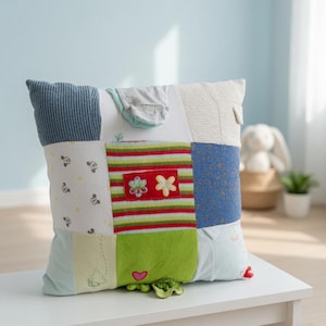Puede incluir: Un cojín cuadrado de patchwork hecho de varios retazos de tela, incluyendo tela blanca con pequeños iconos grises, tela verde con un corazón rojo y un panel central con rayas horizontales rojas, verdes y amarillas con dos aplicaciones de flores.