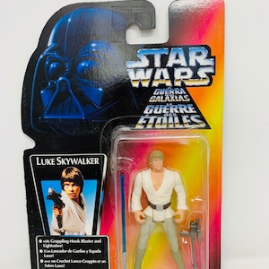 Op de afbeelding: Een plastic action figure van Luke Skywalker uit Star Wars. De figuur draagt een witte tuniek en een bruine broek. Hij houdt een lightsaber en een blaster vast. De verpakking is rood, geel en blauw met het Star Wars-logo en de woorden "Guerra de las Galaxias" en "Guerre des Etoiles".