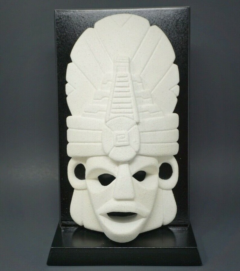 Peut inclure: Masque en c&eacute;ramique blanche avec un visage stylis&eacute; et une coiffe ressemblant &agrave; une pyramide. Le masque est mont&eacute; sur un support en bois noir.