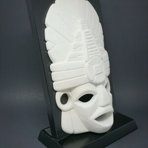 Peut inclure: Masque en c&eacute;ramique blanche avec des d&eacute;tails complexes, mont&eacute; sur un support noir. Le masque pr&eacute;sente un visage stylis&eacute; avec une coiffe.
