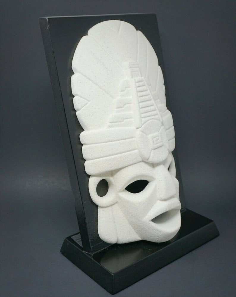 Peut inclure: Masque en c&eacute;ramique blanche avec des d&eacute;tails complexes, mont&eacute; sur un support noir. Le masque pr&eacute;sente un visage stylis&eacute; avec une coiffe.