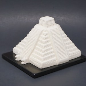 Könnte beinhalten: Weißes Keramikmodell einer Maya-Pyramide mit schwarzem Sockel. Die Pyramide hat ein gestuftes Design mit aufwendigen Details.
