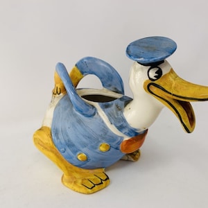 Puede incluir: Una jarra de cerámica con forma de pato de dibujos animados que lleva un sombrero azul. El pato está pintado en tonos de azul, amarillo, naranja y blanco.