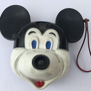 Puede incluir: Una cámara de juguete de plástico en blanco y negro con forma de cabeza de Mickey Mouse. La cámara tiene una correa roja y ojos azules.