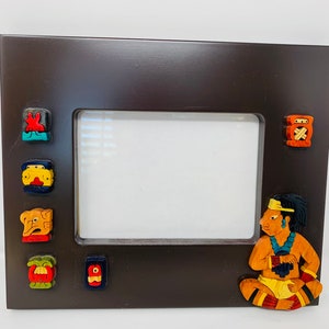 Op de afbeelding: Een donkerbruin houten fotolijst met een witte passe-partout en een gesneden houten figuur van een persoon in traditionele kleding. De lijst is versierd met kleurrijke, gesneden houten figuren rond de opening van de passe-partout.