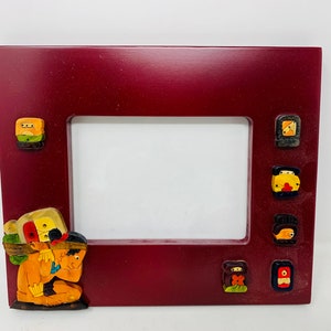 Op de afbeelding: Een rood houten fotolijstje met een decoratieve rand van kleurrijk geschilderde figuren. Het lijstje heeft een rechthoekige opening voor een foto.