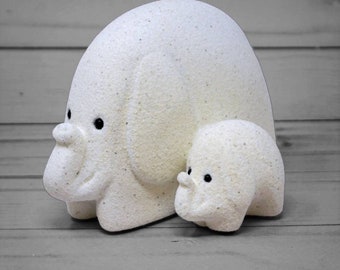 May include: Two white ceramic elephant figurines, one larger than the other, with black eyes. The larger elephant is standing with its trunk curled up, and the smaller elephant is standing behind it with its trunk down.