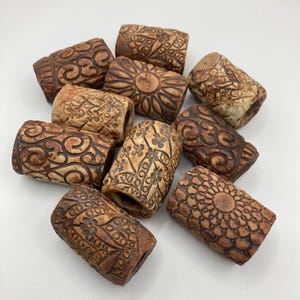 May include: A collection of brown, cylindrical beads with intricate, carved designs. The beads feature floral, geometric, and swirling patterns. The color palette includes shades of brown and tan, with a slightly aged appearance. These beads are likely for jewelry making.