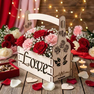 May include: A wooden flower basket with the word "Love" cut out on the front, filled with red roses and pink carnations. The basket has a handle and decorative cutouts of roses and hearts. Scattered around are rose petals, heart-shaped wooden cutouts, and a box of chocolates.