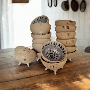 Può includere: Un set di piccole ciotole in ceramica marrone con un design geometrico in bianco e nero. Le ciotole hanno tre gambe e sono disposte su un tavolo di legno.