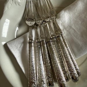 Può includere: Un set di sei forchette d'argento ornate, disposte su un piatto bianco con un tovagliolo di lino bianco. Le forchette presentano dettagliati motivi floreali.