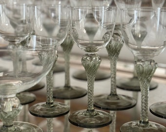 May include: A set of clear glass wine glasses with green stems and bases. The glasses are arranged in a row on a reflective surface.