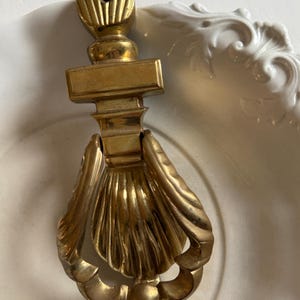 May include: A vintage brass door knocker with a shell design. The knocker features a decorative top with vertical lines and a rectangular midsection. The lower portion is shaped like a scallop shell. The brass has a warm, aged patina.