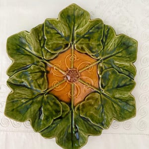 May include: A hexagonal ceramic plate featuring a glossy green leaf-shaped border surrounding a central amber-colored hexagonal section. The center displays a raised, textured brown floral element with six radiating stems extending outward.