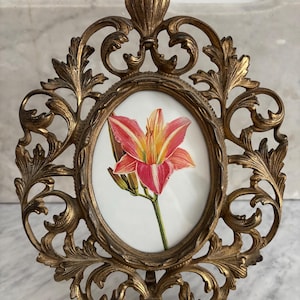 May include: A gold-toned metal picture frame with an ornate design. The frame features a delicate floral pattern and holds a picture of a pink lily.
